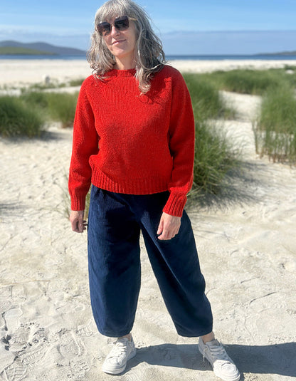 A woman standing on a sandy beach wearing a bright red knit sweater, dark navy wide-leg pants, white sneakers, and sunglasses.