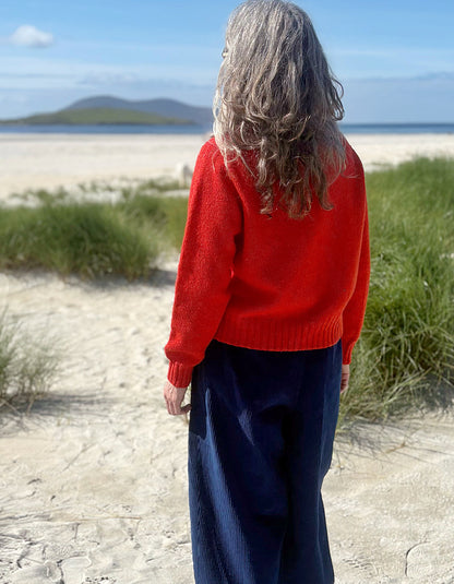 A woman viewed from behind wearing a bright red knit sweater and navy wide-leg trousers, standing on a sandy beach with dune grass and water in the background.