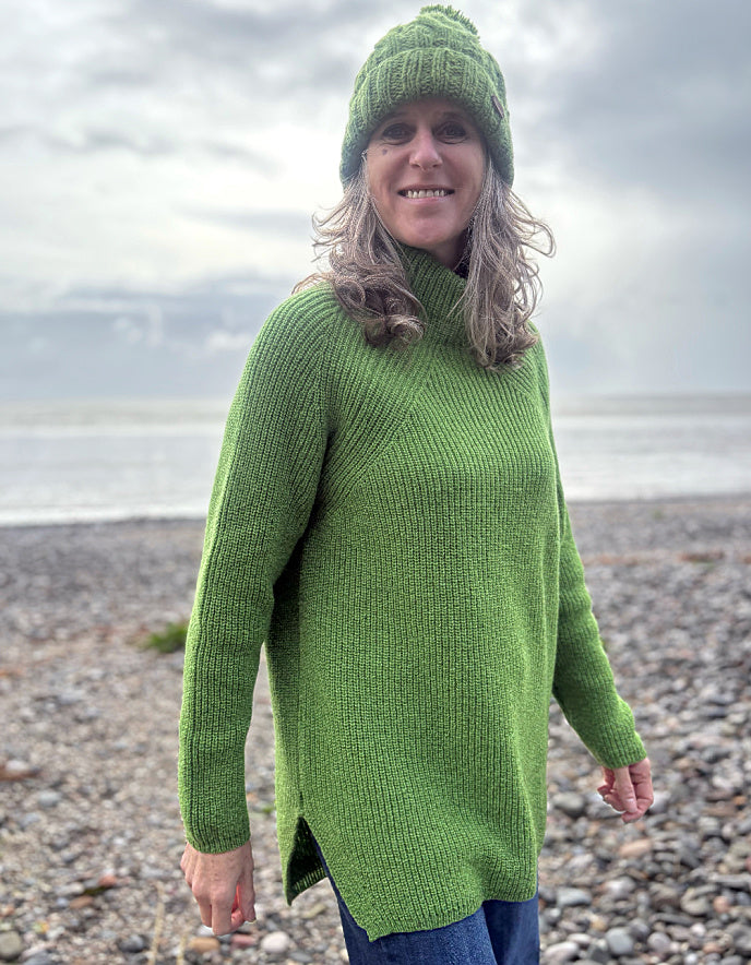 Woman wearing a green knitted sweater and hat standing on a pebbly beach.