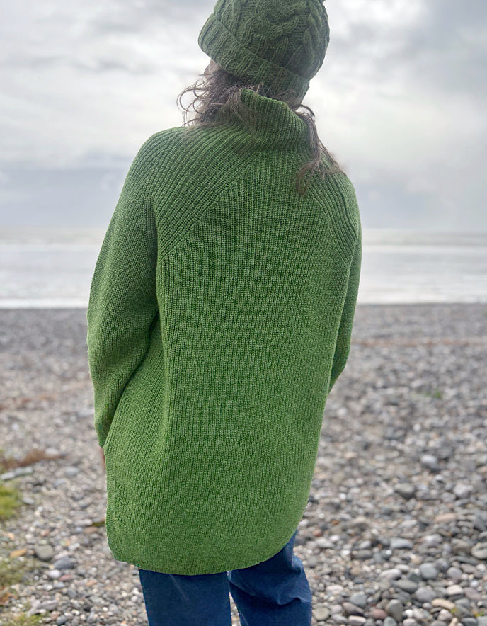Person wearing a green knitted sweater and hat standing on a pebbly beach.