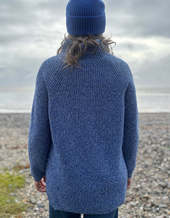 Person wearing a blue knitted sweater and hat standing on a pebbly beach with water in the background