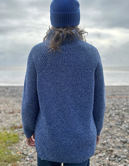 Person wearing a blue knitted sweater and hat standing on a pebbly beach with water in the background