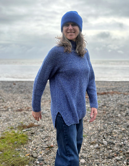 Person wearing a blue sweater and knit hat standing on a pebbly beach.