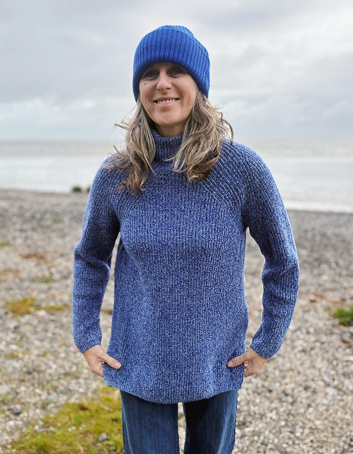 Woman wearing a blue knitted sweater and hat standing on a pebbly beach.