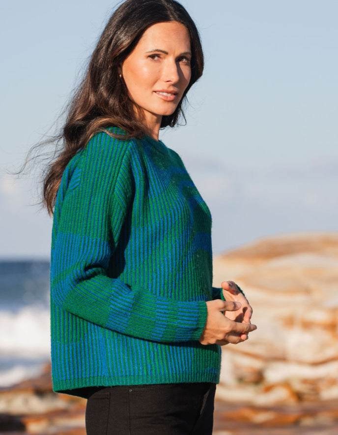 Woman wearing a green and blue sweater standing on a beach with ocean and sand in the background.