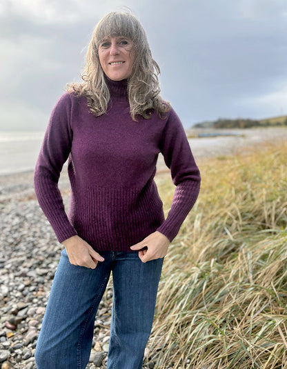 Woman wearing a purple sweater and blue jeans standing on a beach.