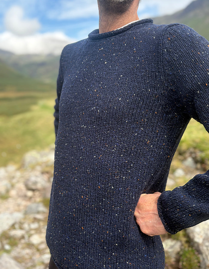 A close-up of a man wearing a dark navy blue speckled knit sweater, standing outdoors with a blurred mountain and grassy landscape in the background.
