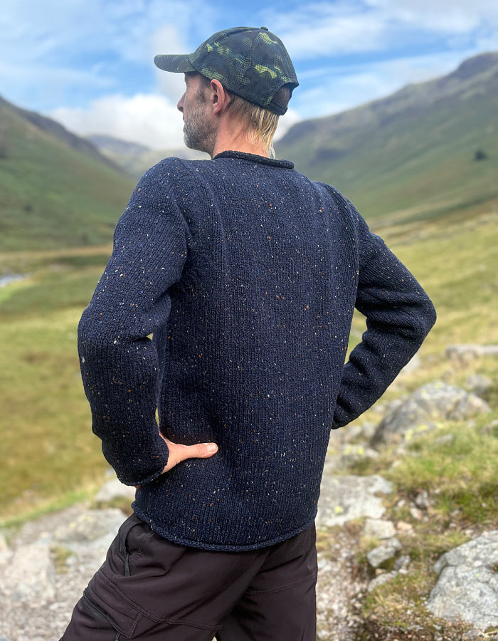 A man wearing a dark navy speckled knit sweater and a camouflage cap, standing outdoors in a mountain landscape and looking away from the camera.