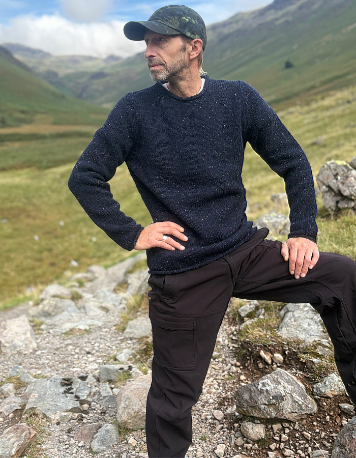 A man wearing a dark navy speckled knit sweater and dark brown pants stands outdoors on a rocky path with a mountain landscape in the background.