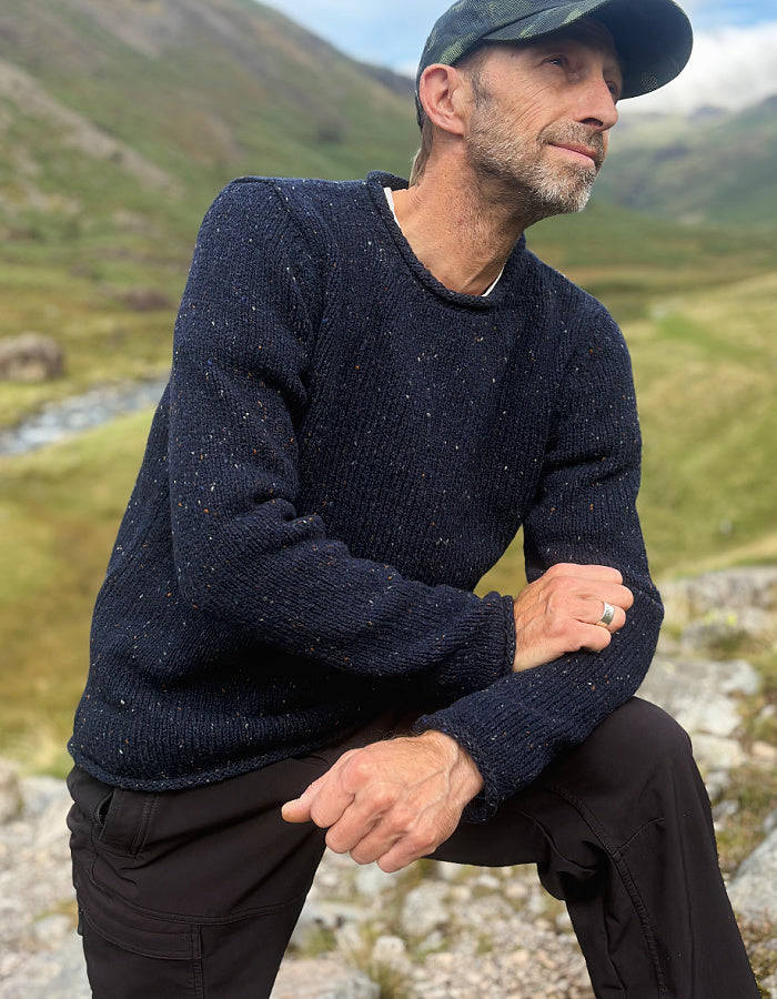 A man wearing a dark navy speckled knit sweater and a dark cap, crouching outdoors in a grassy mountain landscape.