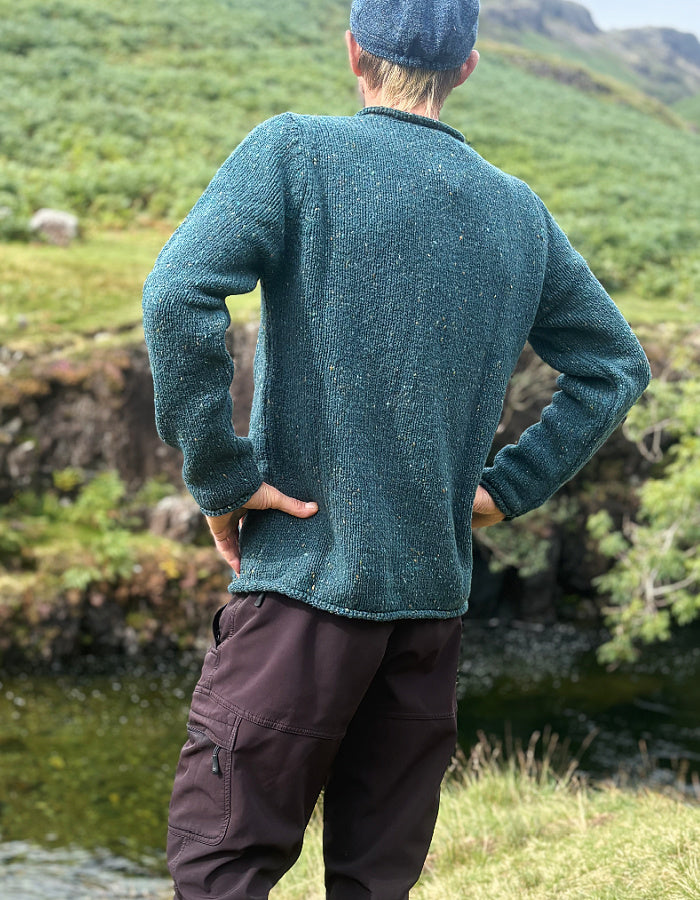 A man wearing a textured teal-green knit sweater and dark brown pants, standing outdoors with his back to the camera near a grassy riverbank.