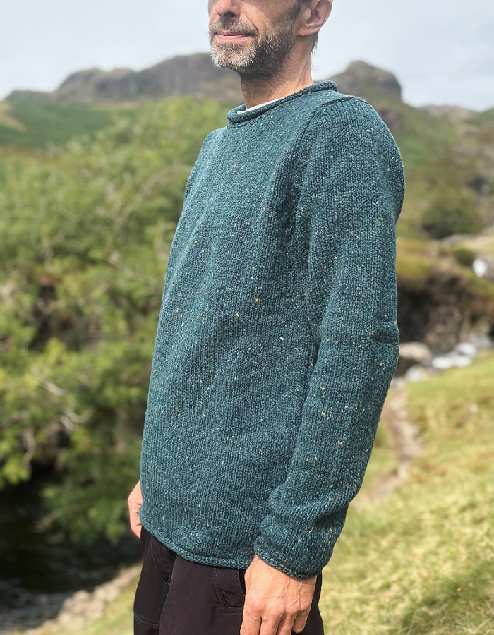 A man wearing a textured, speckled teal-green knit sweater, standing outdoors in a grassy, rocky landscape.