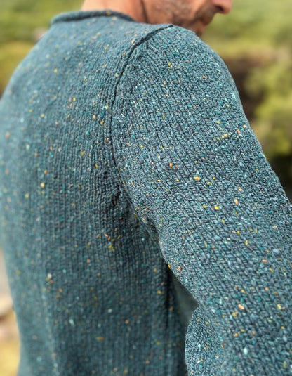 Close-up of a person wearing a textured knit sweater in a heathered teal blue-green color with small multicolored flecks, photographed outdoors with a blurred natural background.
