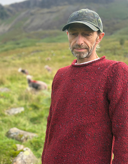 A man wearing a textured red knit sweater and a camouflage baseball cap stands outdoors in a grassy mountain landscape with sheep in the background.