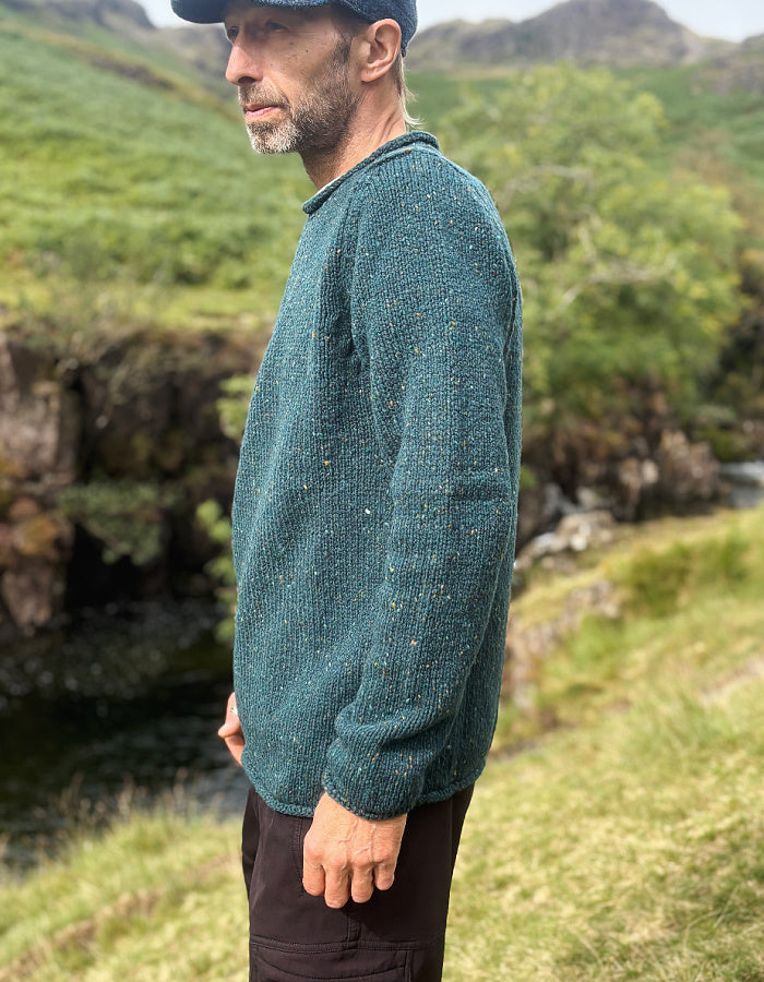 A man wearing a textured teal-green knit sweater and dark trousers stands outdoors in a grassy, rocky landscape.