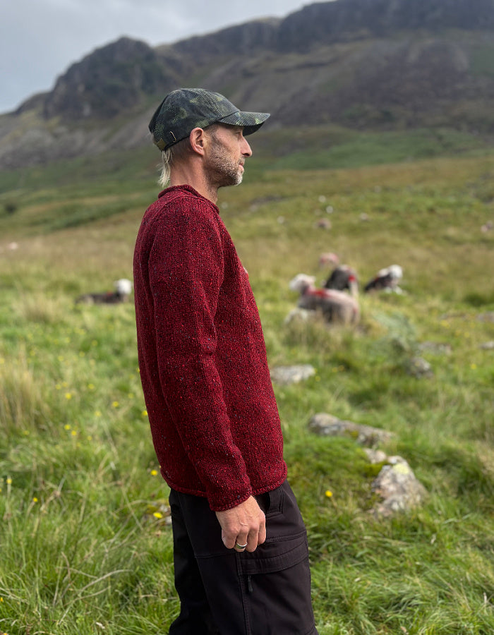 A man wearing a textured dark red knit sweater, black trousers, and a camouflage cap stands in profile in a grassy mountain landscape.
