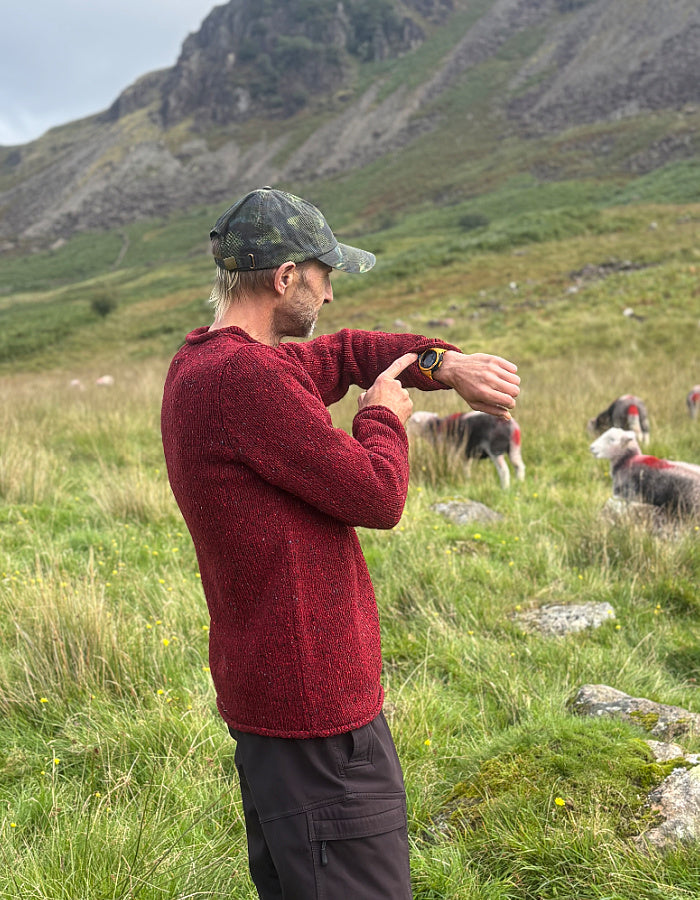 A man in a red knit sweater and camouflage cap stands in a grassy mountain field, looking at a smartwatch. Sheep are visible in the background under an overcast sky.