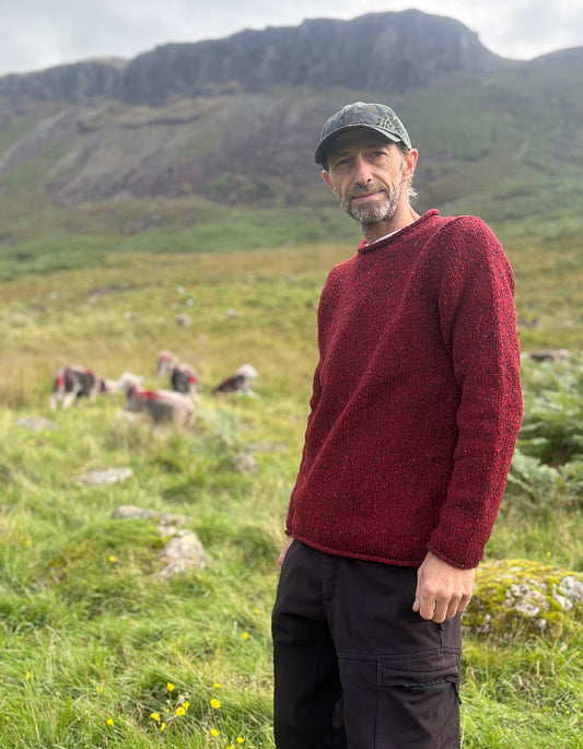 A man wearing a textured dark red knit sweater, black cargo pants, and a camouflage baseball cap stands in a grassy mountain meadow with sheep in the background.