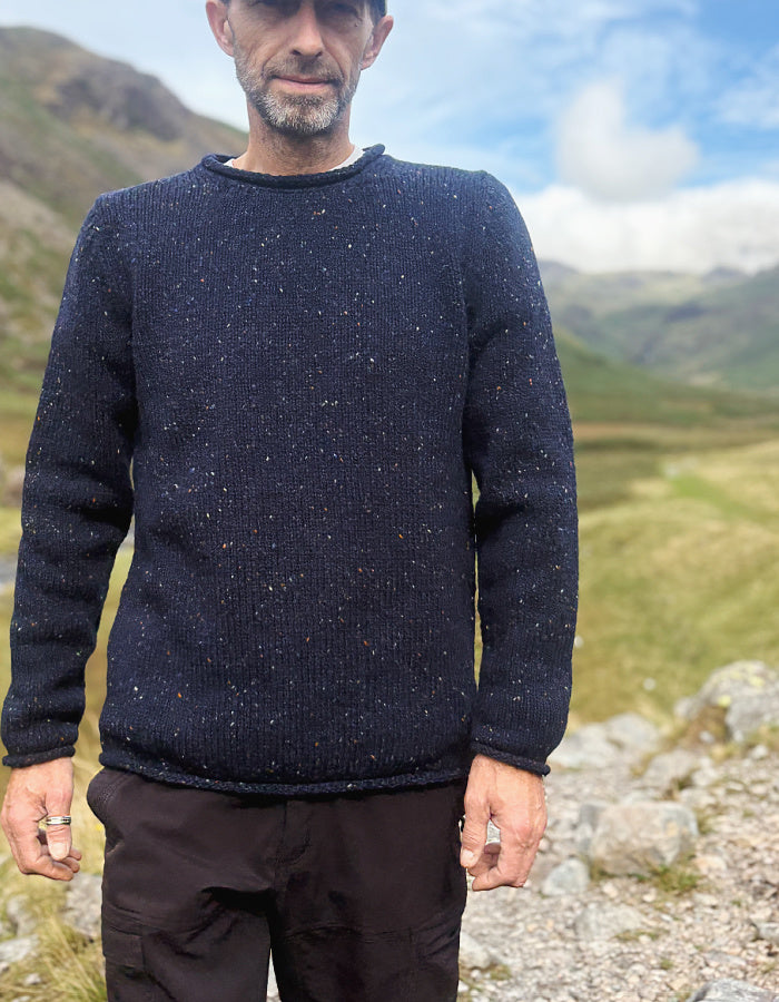 A man wearing a dark navy speckled knit sweater and dark trousers, standing outdoors in a mountain valley landscape.