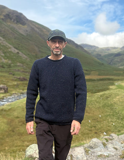 A man wearing a dark navy speckled knit sweater, dark trousers, and a dark cap stands outdoors in a grassy mountain valley with a stream and hills in the background.