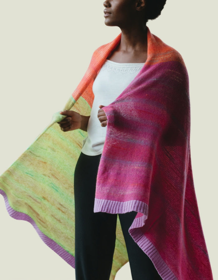 A woman wearing a large, multicolored knit shawl draped over her shoulders, shown from the side against a plain light beige background.