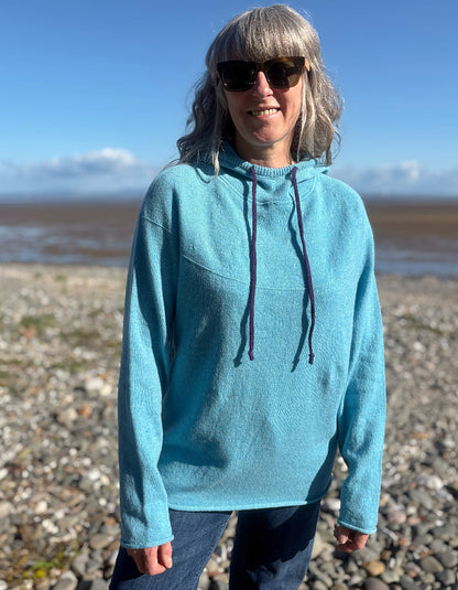 A woman with gray hair and sunglasses wearing a light blue knit hoodie sweater, standing on a pebble beach with water and sky in the background.