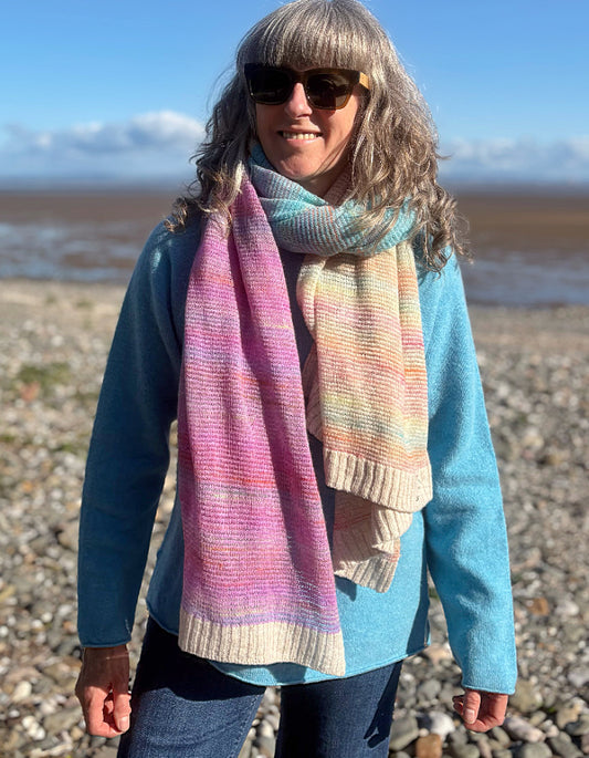A woman standing on a pebble beach wearing a pastel striped knit scarf with pink, blue, yellow, and cream tones, paired with a light blue sweater and dark sunglasses.
