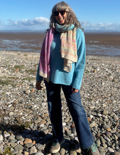 A woman stands on a rocky beach wearing a turquoise knit sweater, a multicolor striped knit scarf, dark blue jeans, and sunglasses, with water and sky in the background.
