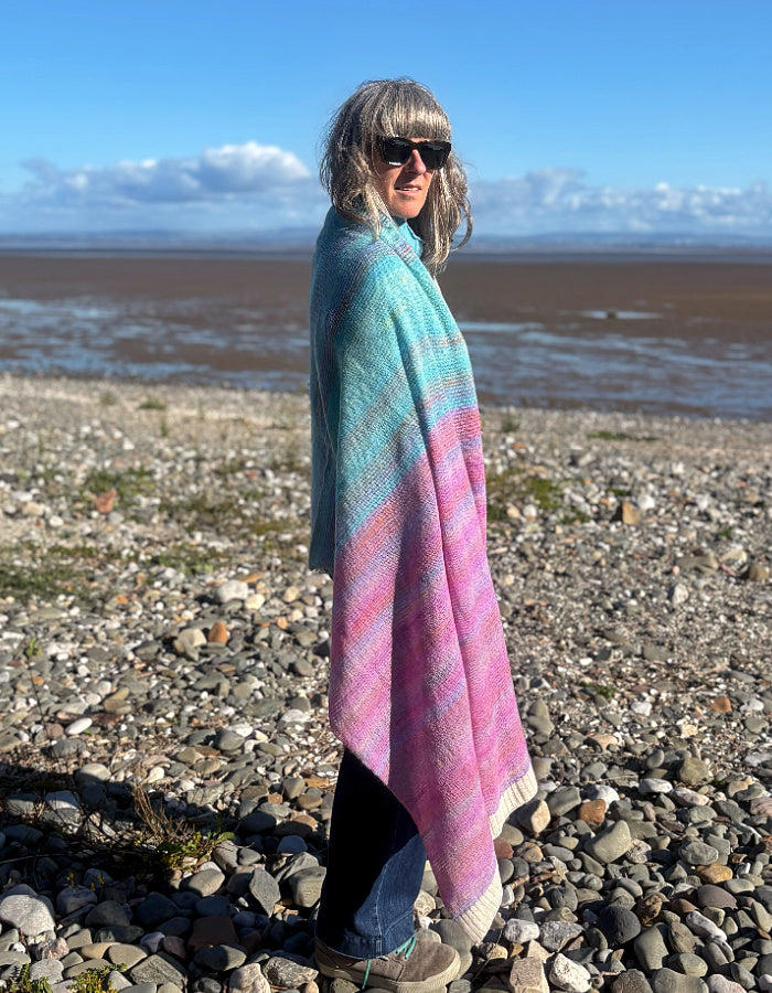 A woman wearing sunglasses stands on a rocky beach, wrapped in a soft, striped knit shawl with wavy bands of turquoise, pink, and lavender. The background shows a wide expanse of wet sand and distant water under a blue sky.