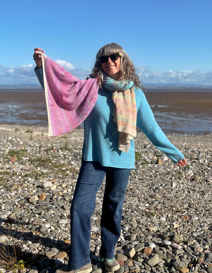 A woman standing on a rocky beach wearing a light blue sweater, dark blue jeans, and a striped knit scarf, while holding up a pink and purple knitted shawl or wrap in one arm against a blue sky and water background.