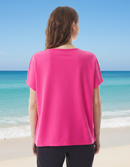 Person wearing a pink short sleeve t shirt standing on a beach with clear blue water and sky.