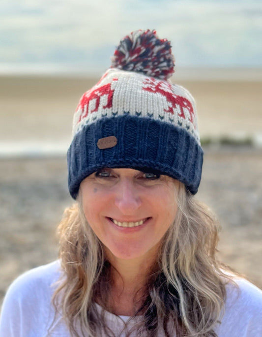 A woman smiling and wearing a knitted beanie with a reindeer pattern and a multicolored pom-pom.