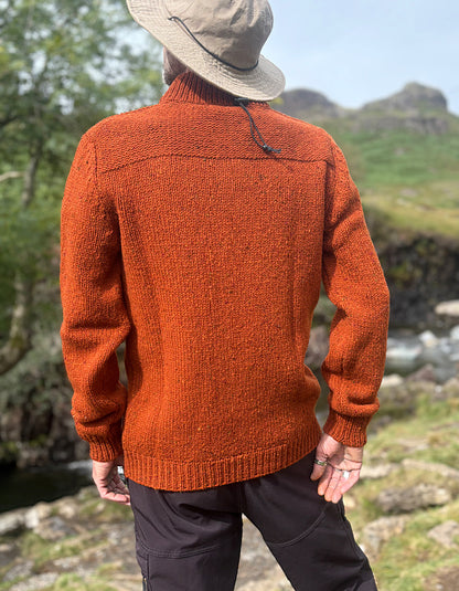 A man wearing a textured rust-orange knit sweater, a beige wide-brim hat, and dark trousers, photographed from behind in an outdoor mountain landscape.
