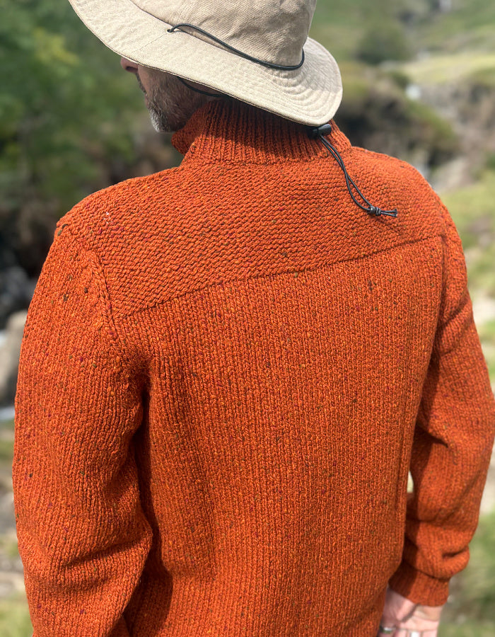 A man wearing a textured orange knit turtleneck sweater, a beige wide-brim hat, and a beanie, photographed from behind in an outdoor setting.