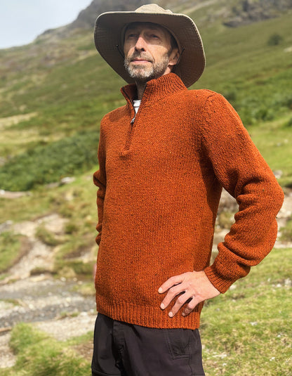 A man wearing a textured rust-orange quarter-zip sweater, a beige wide-brim hat, and dark trousers stands outdoors in a grassy mountain landscape.