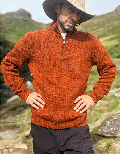 A man wearing a textured rust-orange quarter-zip sweater, a beige wide-brim hat, and dark cargo pants, standing outdoors in a grassy mountain setting.
