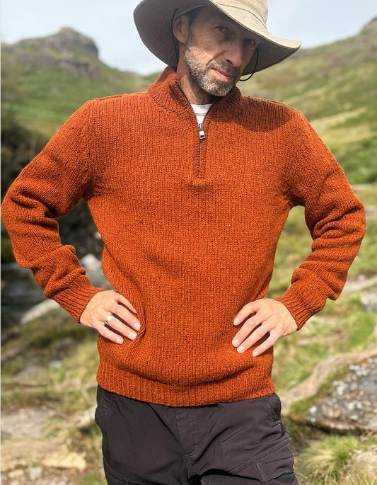 A man wearing a textured rust-orange quarter-zip sweater, a beige wide-brim hat, and dark cargo pants, standing outdoors in a grassy mountain setting.