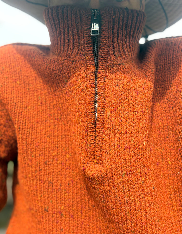 Close-up of a bright orange knit quarter-zip sweater with a ribbed mock neck collar and a black zipper, worn by a person wearing a wide-brim hat.