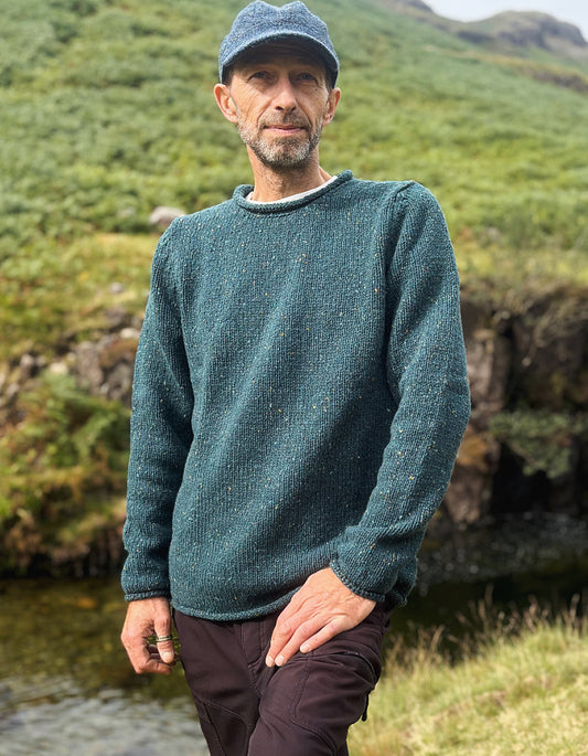 A man wearing a textured teal-green crewneck sweater and a matching flat cap, standing outdoors in a grassy, rocky mountain environment.