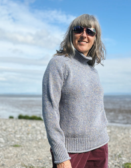 Person wearing a gray sweater and sunglasses on a beach with a blue sky.