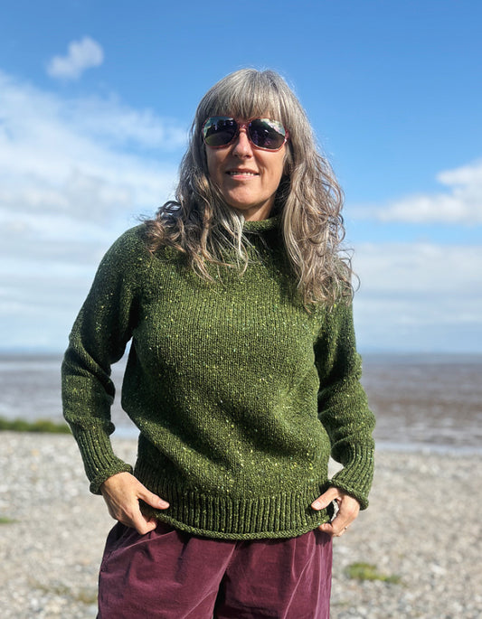 Person wearing a green donegal tweed turtle neck  sweater and sunglasses on a beach with a blue sky.