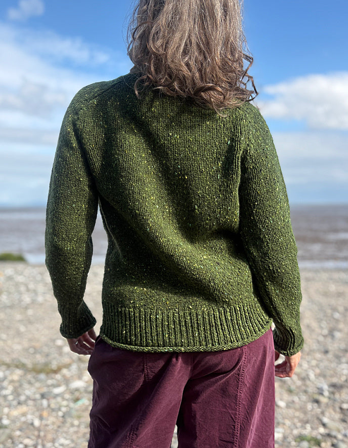Person wearing a green donegal tweed turtle neck  sweater and sunglasses on a beach with a blue sky.
