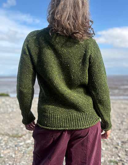 Person wearing a green donegal tweed turtle neck  sweater and sunglasses on a beach with a blue sky.