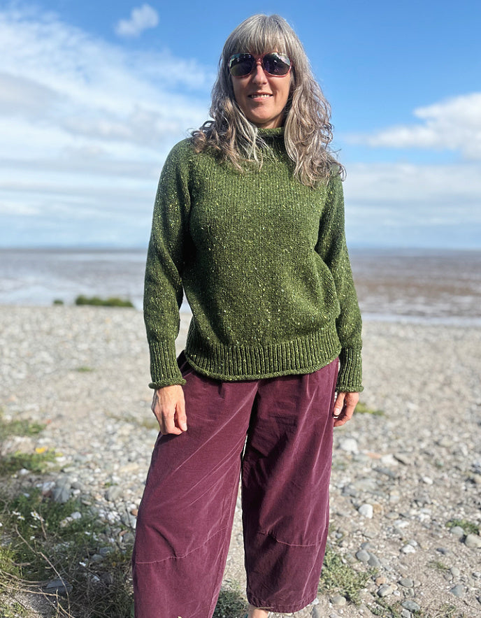 Person wearing a green donegal tweed turtle neck  sweater and sunglasses on a beach with a blue sky.