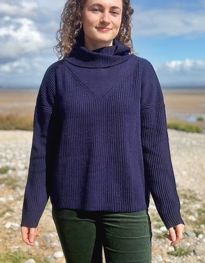 A woman wearing a dark navy blue ribbed turtleneck sweater and dark green pants, standing outdoors on a rocky shoreline with water and a cloudy sky in the background.