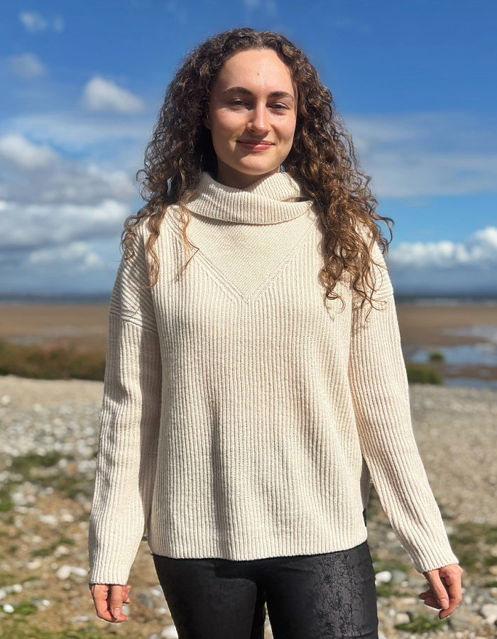 A woman with long curly brown hair stands outdoors on a rocky shoreline under a blue sky, wearing a cream ribbed cowl-neck sweater and black pants.