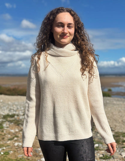 A woman with long curly brown hair stands outdoors on a rocky shoreline under a blue sky, wearing a cream ribbed cowl-neck sweater and black pants.