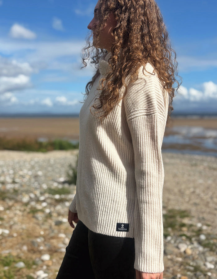 A woman with long curly brown hair stands outdoors in profile wearing a cream ribbed knit sweater and black pants, with a rocky shoreline and blue sky in the background.