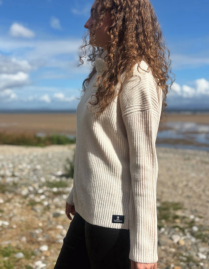 A woman with long curly brown hair stands outdoors in profile wearing a cream ribbed knit sweater and black pants, with a rocky shoreline and blue sky in the background.