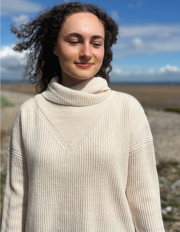 A woman with curly dark hair wearing a cream-colored ribbed turtleneck sweater, standing outdoors against a blurred background of blue sky and water.
